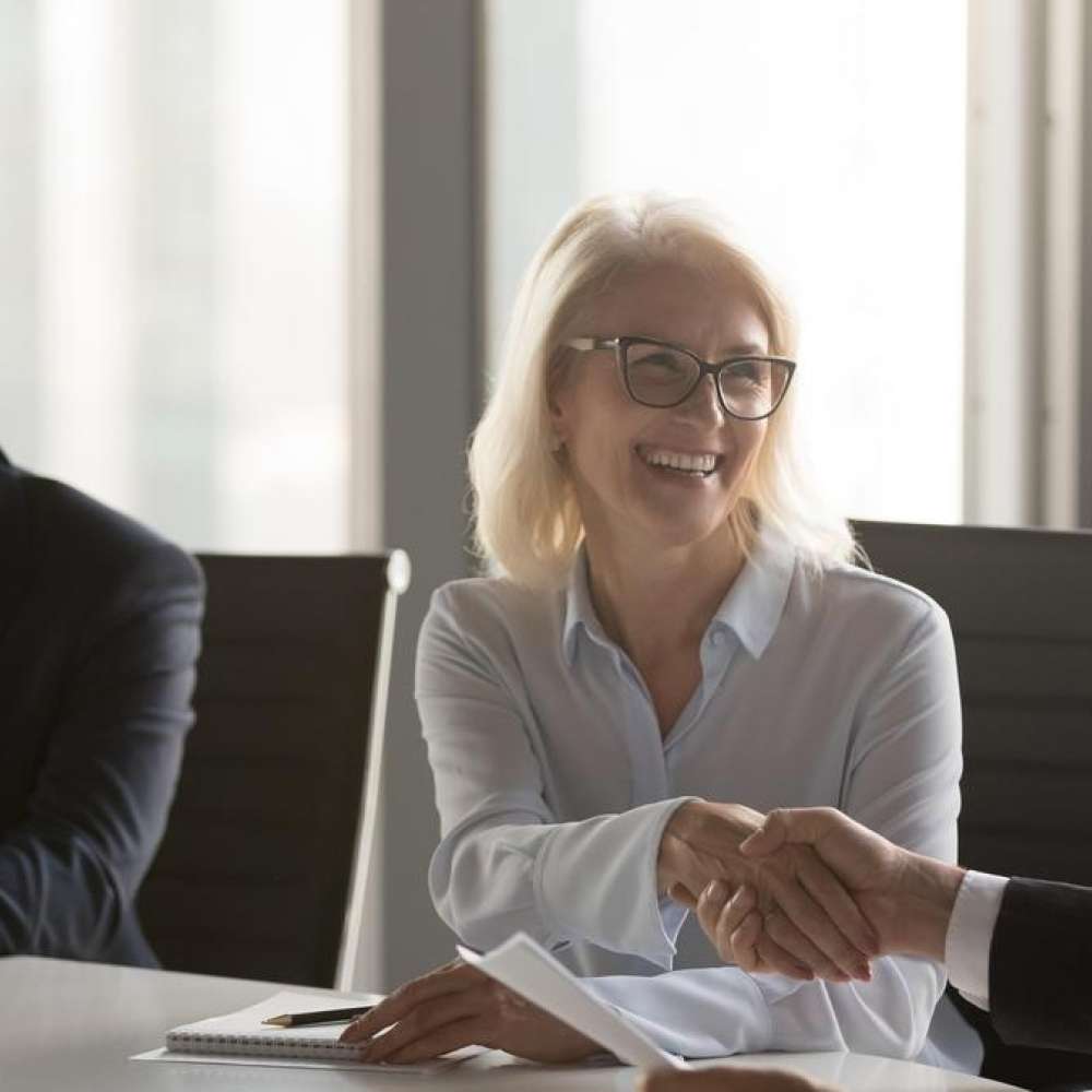 Two people are sitting at a table in an office shaking hands.